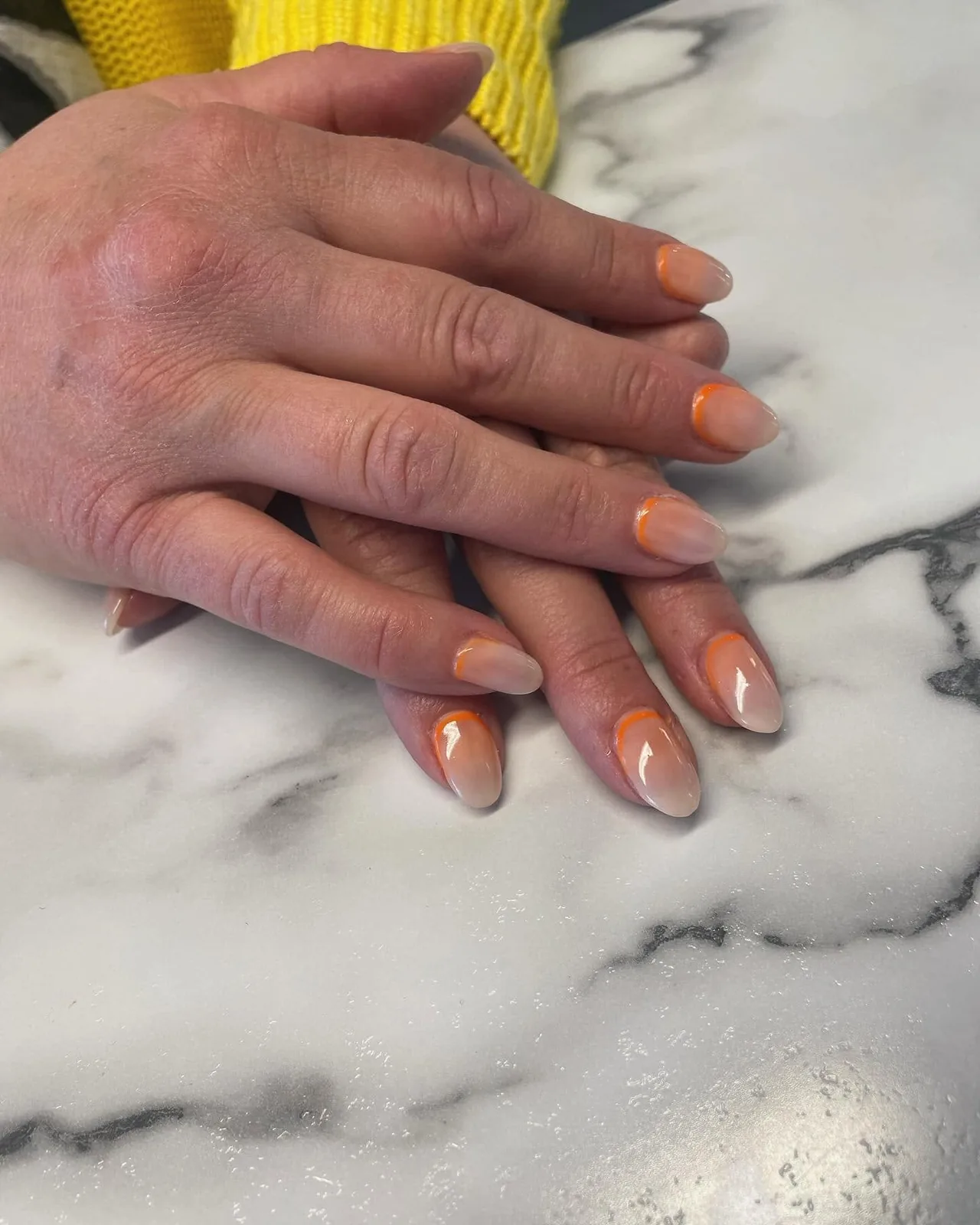 Mains sur un comptoir de marbre, ongles nude en dégradé soulignés d’un fin liseré orange corail.