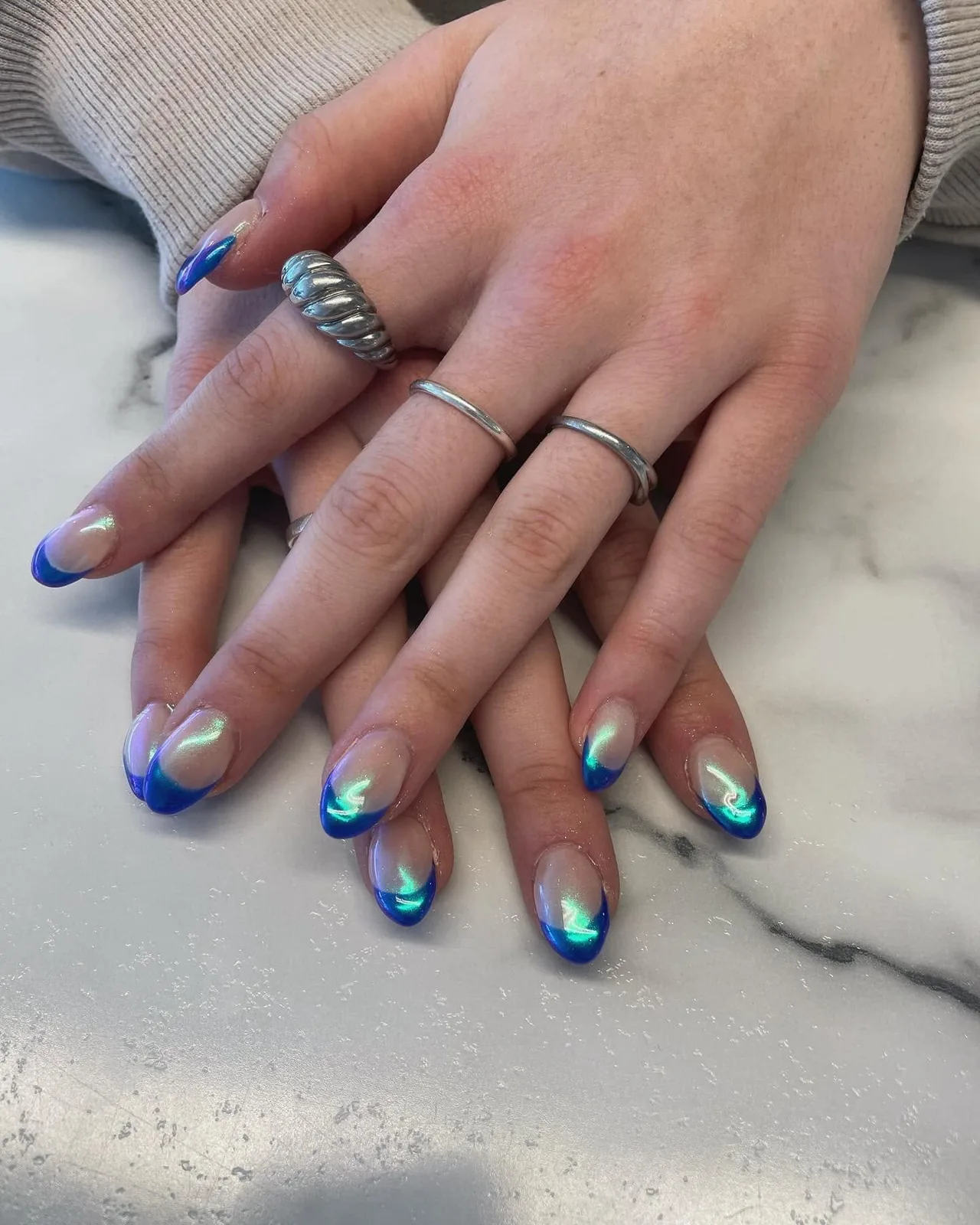 Mains baguées posées sur du marbre, ongles amande à french bleu franc avec un reflet vert irisé.