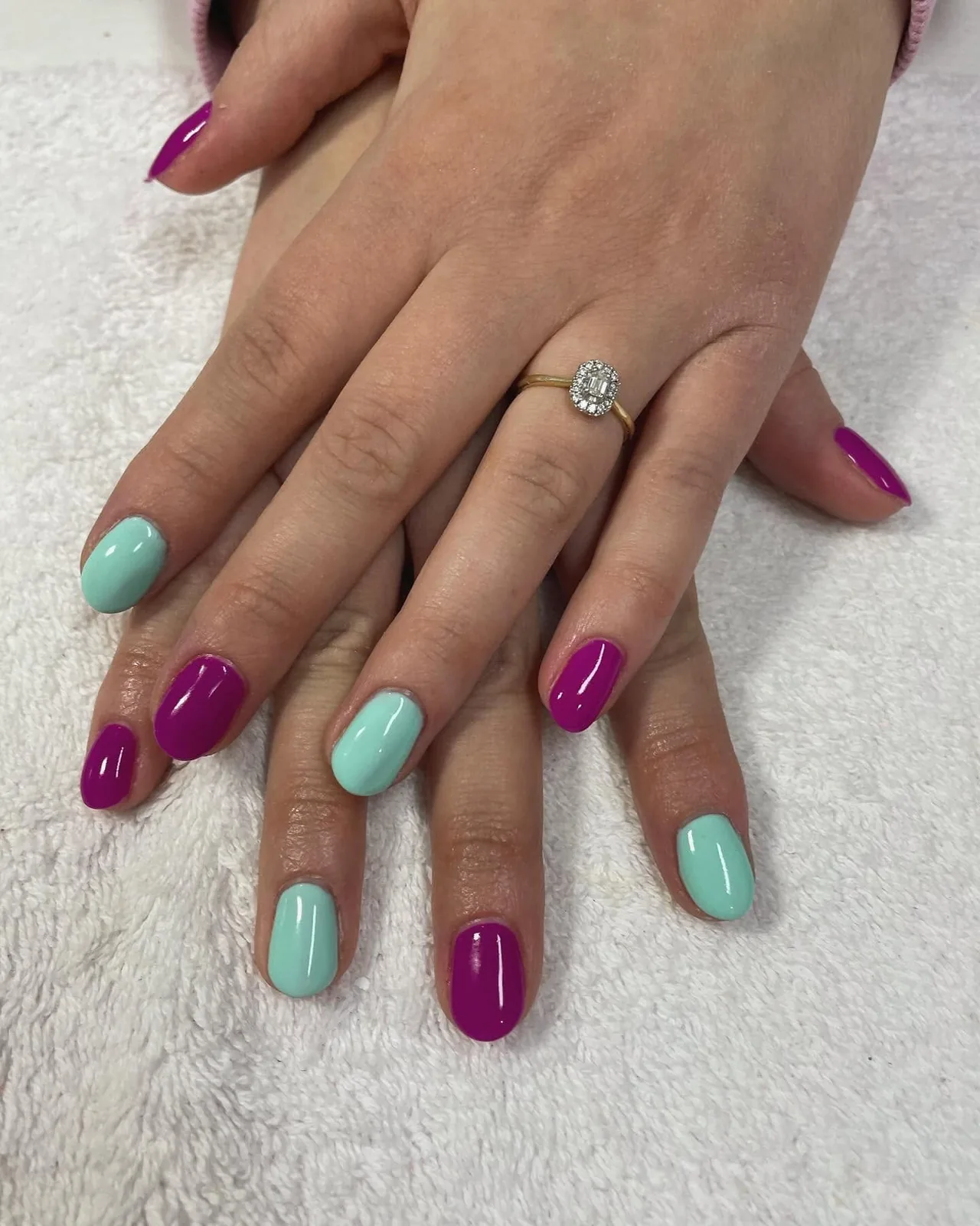 Mains posées sur une serviette blanche, ongles courts alternant vernis magenta et vernis vert menthe.